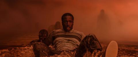 A terrified Black man sits in a foggy orange landscape, with looming shadowy figures in the background