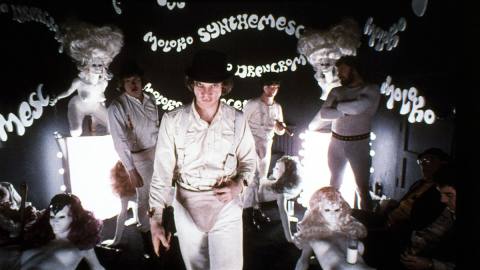Malcolm McDowell as Alex in A Clockwork Orange stands in a darkened milk bar full of naked female mannequins, with fictional drug names all over the walls