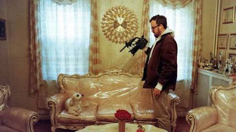 John Wilson stands with a camera filming a dog sitting on nice furniture covered with plastic wrap 