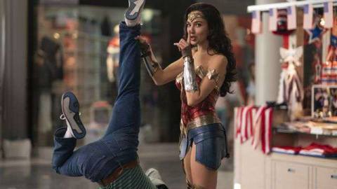 Gal Gadot as Wonder Woman holds a man upside down by his ankle while holding a finger to her lips in a “shhh” gesture in Wonder Woman 1984. 
