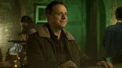 Alan Tudyk sits as a bar wearing a weird smile as a disguised alien in Resident Alien