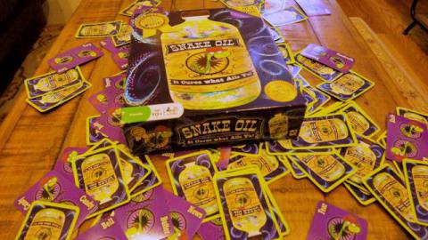 The box to the board game Snake Oil sits on a pile of cards from the game.