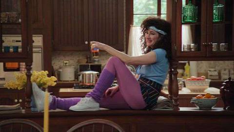 Agnes, wearing ‘80s workout gear, sits on a countertop.