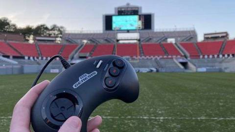 a hand holds a Sega Genesis gamepad with a large stadium video board far in the distance