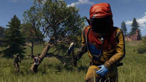 a man in a hazmat suit with a red hood brandishes an ax as two people chop down a tree behind him in Rust Console Edition
