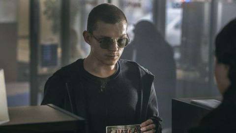 Tom Holland as Cherry with a shaved head standing at a bank teller window with a dollar bill that says “I have a gun” in red marker