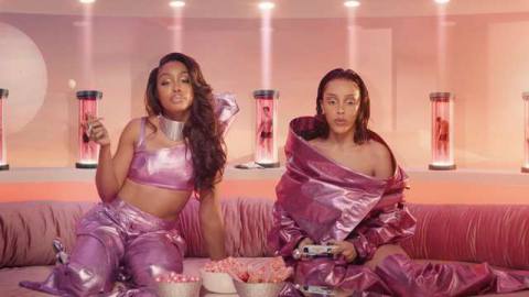 Doja Cat and SZA sitting on a couch, wearing shiny pink outfits while playing video games .
