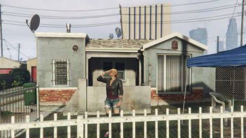 Grand Theft Auto Online - a blonde woman in jean shorts and a leather jacket facepalms. She is standing in front of a blue and brick house.