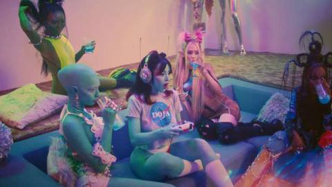 Doja Cat sits in a futuristic midcentury style sunken living room while holding on a playstation 5 controller. she’s surrounded by a ton of alien girls
