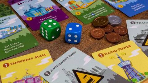 A pile of bits from Machi Koro, including a shopping mall, an amusement park, and a radio tower. Two dice and some coins sit in the center of the frame.