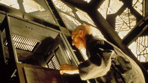 A bald man approaches an aperture in a grid-based wall in the science fiction movie Cube 