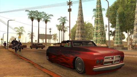 Grand Theft Auto: San Andreas - a red and black car speeds down a Los Angeles road, pursued by attackers on foot