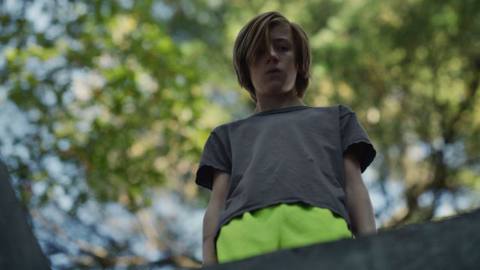 13-year-old John, with longer hair and neon yellow shorts, stands over a hole with the camera looking up at him