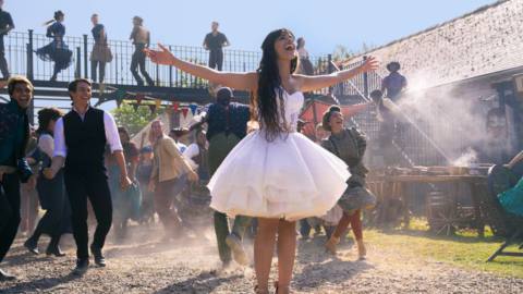 Camila Cabello, in a thigh-length floofy dress, sings outdoors in front of a parade of bland smiling people in Amazon’s Cinderella.