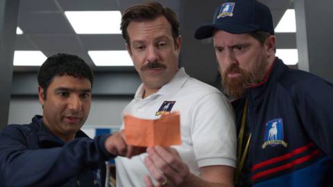 Ted Lasso and his assistant coaches lean over a note