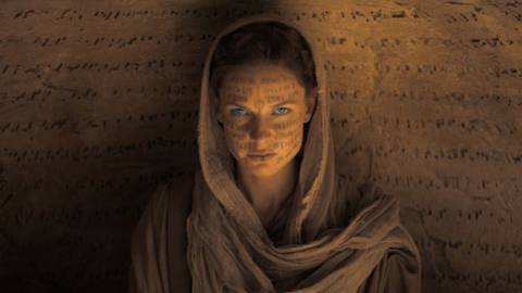 Rebecca Ferguson, in sandy robes, sits in front of a wall carved with lines of alien text. Her face is also scrawled with alien text, and her eyes are glowing blue in Dune. 