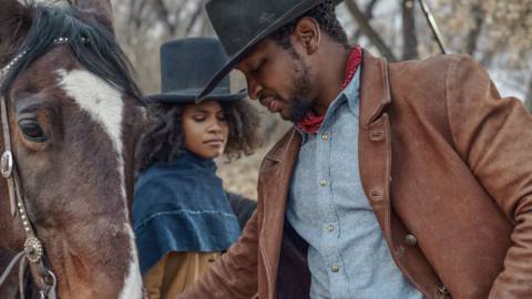 Zazie Beetz and Jonathan Majors wear cowboy hats and stand by a horse, pointedly not looking at each other, in The Harder The Fall