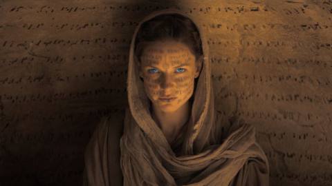 Rebecca Ferguson, in sandy robes, sits in front of a wall carved with lines of alien text. Her face is also scrawled with alien text, and her eyes are glowing blue in Dune.