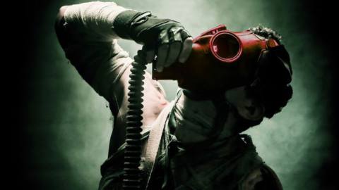 A bandaged man looks up, into the light, his hand poised to remove his red gas mask.