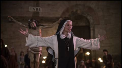 Virginie Efira as ‘Benedetta’ standing in front of a crucifix with her arms outstretched in Paul Verhoeven’s ‘BENEDETTA.’