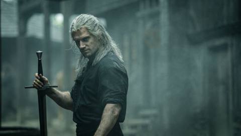 Geralt of Rivia, a brawny gray-haired man, holds a sword upside down by the hilt in The Witcher