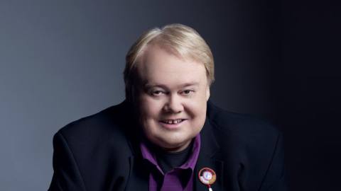 A portrait of comedian Louie Anderson, seated on a stool and smiling.