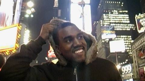 a grainy video screenshot from jeen-yuhs: A Kanye Trilogy of Kanye West as a young man, wearing a black winter coat with a fur-lined hood, smiling as he stands in Times Square