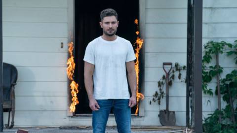 Zac Efron in Firestarter, standing outside in front of a house with an open, burning doorway