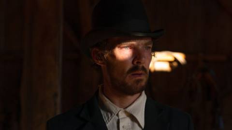 Benedict Cumberbatch in suit and hat in The Power of the Dog, standing in a dark room with bright light falling on one eye