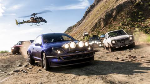 Six cars (four in the foreground, two in the background) race along a wide and mountainous road. It is daytime and the skies are clear with faint wisps of clouds. A news-reporter helicopter hovers above the road following the racing action.