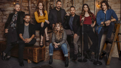 The cast of Critical Role pictured against a rough-hewn wooden wall.