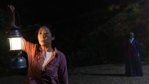 Sandra Oh holds up a lantern outside on a dark beach while a weird figure looms behind her in Umma