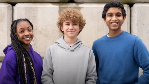 a girl and two boys stand smiling. the girl has long hair in braids and wears a purple hoodie. the middle boy has fluffy blonde hair and smirks. the boy on the right is taller than the other two and wears a blue long-sleeve shirt. they play annabeth, percy, and grover