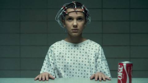Eleven (played by Millie Bobby Brown) sits at a table staring straight ahead while wearing a cap of wires on her head. A can of coke sits on the table to the left of her.