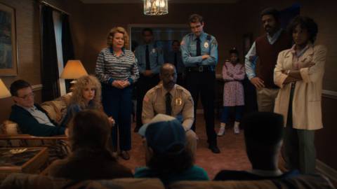 Three of the Stranger Things kids sitting with their back to the camera, with parents and cops looking at them