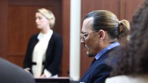 Amber Heard stands in the background of the image, testifying, and Johnny Depp sits in the foreground, at the defamation trial
