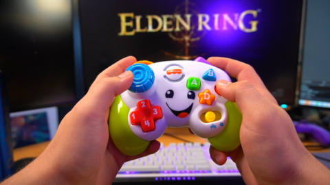 a photo of two hands holding a fisher-price toy controller meant for young kids. the hands are in front of the loading screen for elden ring.