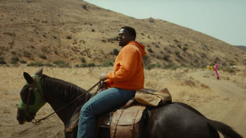 Daniel Kaluuya in Jordan Peele’s Nope. He’s wearing a brightly colored hoodie and jeans, riding a dark horse through a bare and mountainous desert landscape. In the distance there is one flopping Air Dancer machine. 