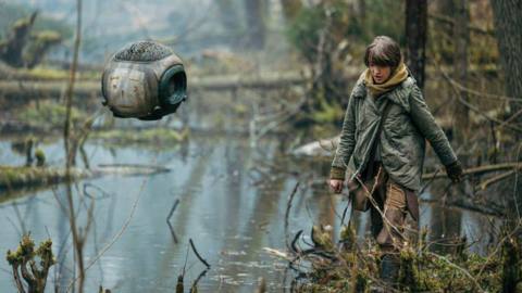 Raffiella Chapman wanders through marshy turf in a post-apocalyptic Earth, with a floating robot companion, in Vesper