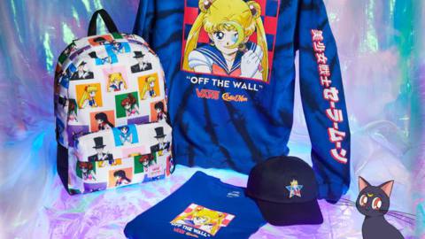 A staged photograph with a tie-dye Sailor Moon hoodie, T-shirt, ball cap, and backpack. There’s an illustration of Sailor Moon’s cat Luna, in the bottom right corner of the image.