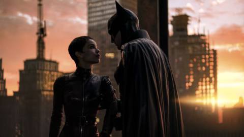Left to right: Zoë Kravitz as Selina Kyle/Catwoman and Robert Pattinson as Bruce Wayne/Batman in The Batman. He grips her arm and they stare into each others’ eyes on a rooftop as the sun rises over the skyline behind them.