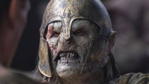 A close-up of an orc in The Lord of the Rings: The Rings of Power. The creature has sallow skin and is wearing a rusted elven helmet as it snarls.