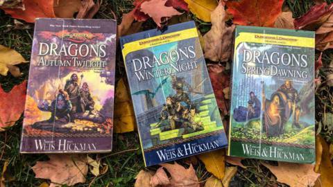 Three books on a bed of fallen leaves. Titled Dragons of Autumn Twilight, of Winter Night, and of Spring Dawning.