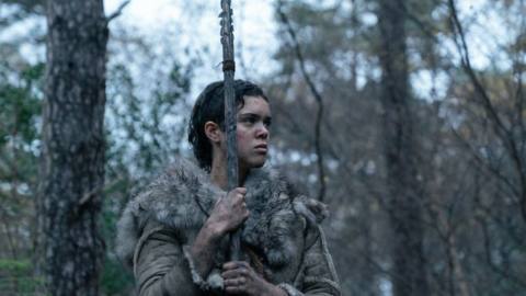 A prehistoric woman, Beyah (Safia Oakley-Green), dressed in furs and holding a spear, stands in the forest in Out of Darkness
