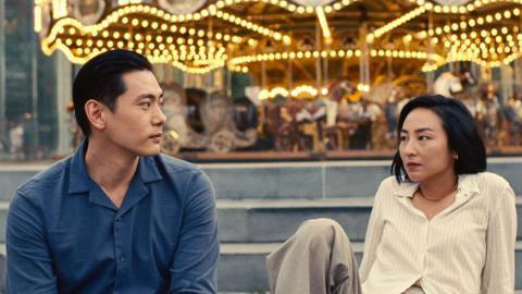 Greta Lee and Teo Yoo look at each other longingly in front of a carousel in Past Lives