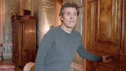 Willem DaFoe reaches for the handle of a secret door on the set of Poor Things
