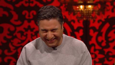 Nick Mohammed laughs on the Taskmaster stage, with a red and black background behind him