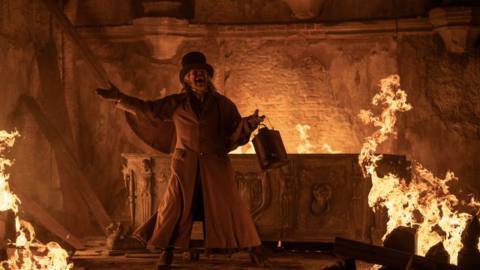 Willem Defoe holding a lantern and standing by an altar with fire all around him in Nosferatu