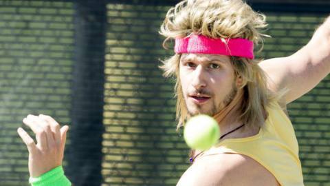 Andy Samberg, with long blond hair, a pink headband and yellow muscle shirt, winds up to hit a tennis ball in 7 Days in Hell
