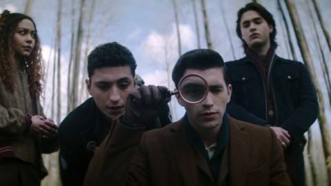 LtR: Kassius Nelson as Crystal Palace, Jayden Revri as Charles Rowland, George Rexstrew as Edwin Payne, and Joshua Colley as Monty in Dead Boy Detectives. Crystal and Monty look skeptical as Charles and Edwin crouch down closer to the camera on the ground. Edwin examines something someone comically with a large magnifying glass held over one eye. 